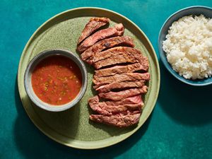 Crying Tiger (Thai-style grilled steak) on a green ceramic plate, with a small ceramic bowl holding the dry chile dipping sauce). On the periphery is a small bowl of white rice.