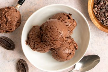 四勺巧克力冰淇淋放在一个白色的圆形陶瓷碗里。碗的外围是一个金属勺子，一个装着可可粒的小木碗，两块巧克力，和一个装着冰淇淋的勺子。