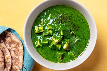 Bright green Palak Paneer in a white bowl one an orange back next to some roti