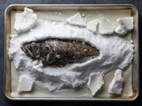 A whole roasted black bass that was buried in a mound of salt on a baking sheet is exposed after the salt crust was cracked and removed.