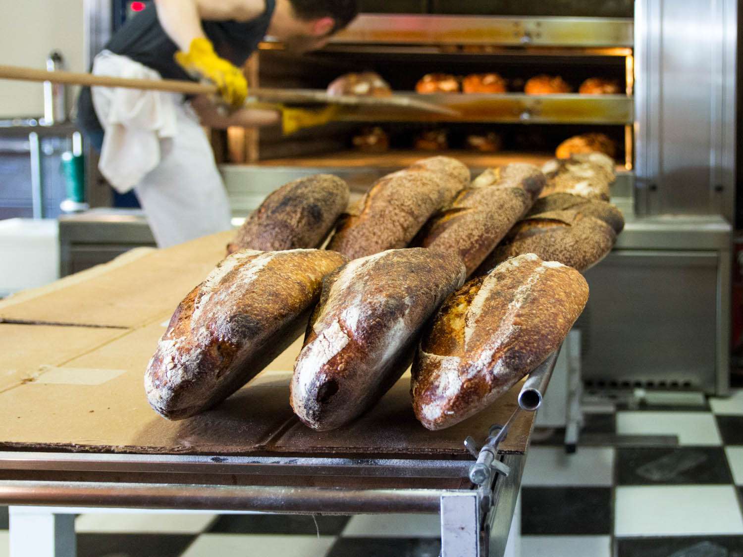 塔廷面包店厨房里面包师桌子上的面包。在背景中，有人正在用一个长柄削皮来移动商用烤箱中的面包。