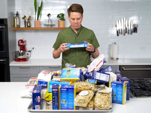 萨莎马克思从严肃的站在厨房吃面条放在柜台上的一堆不同的包在他的面前。