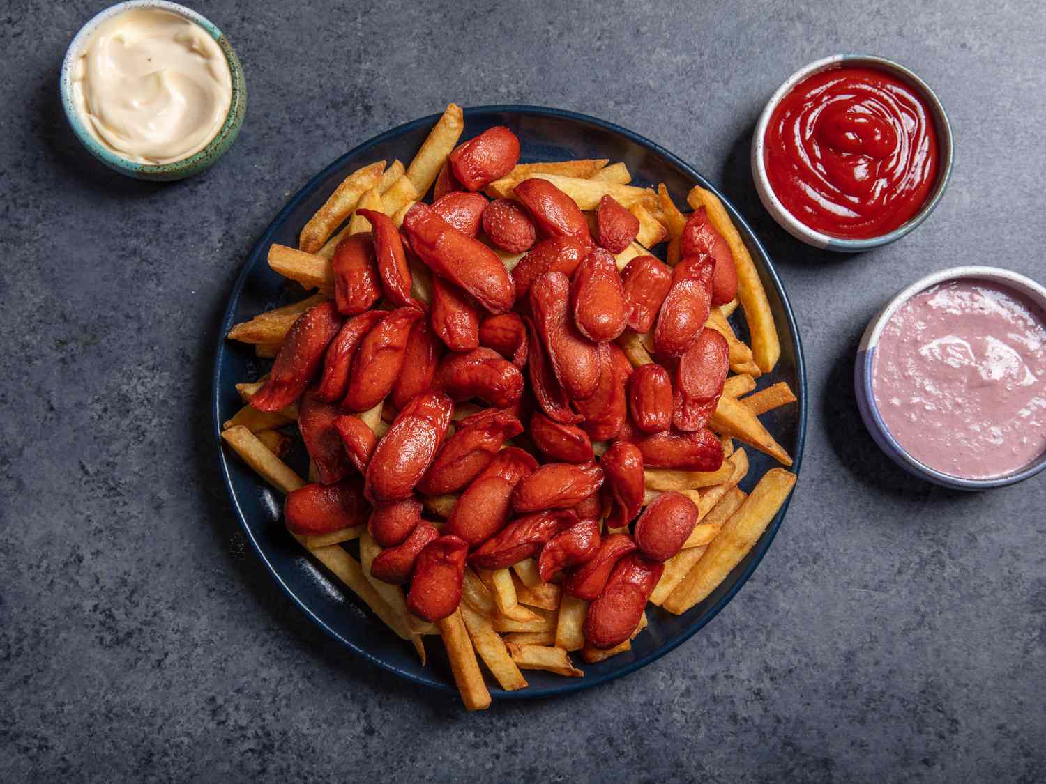 A large platter of salchipapa (french fries with sliced hot dogs piled on top) and a few dipping sauces alongside