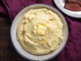 A bowl of fluffy mashed potatoes sits on a table. It's topped with a pat of melting butter and a grinding of fresh black pepper.