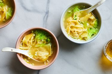 Two bowls of wonton soup on a marble surface. Each bowl has a white ceramic soup spoon in it. There is another bowl on the top left corner of the image, and a glass of liquid on the bottom right periphery.