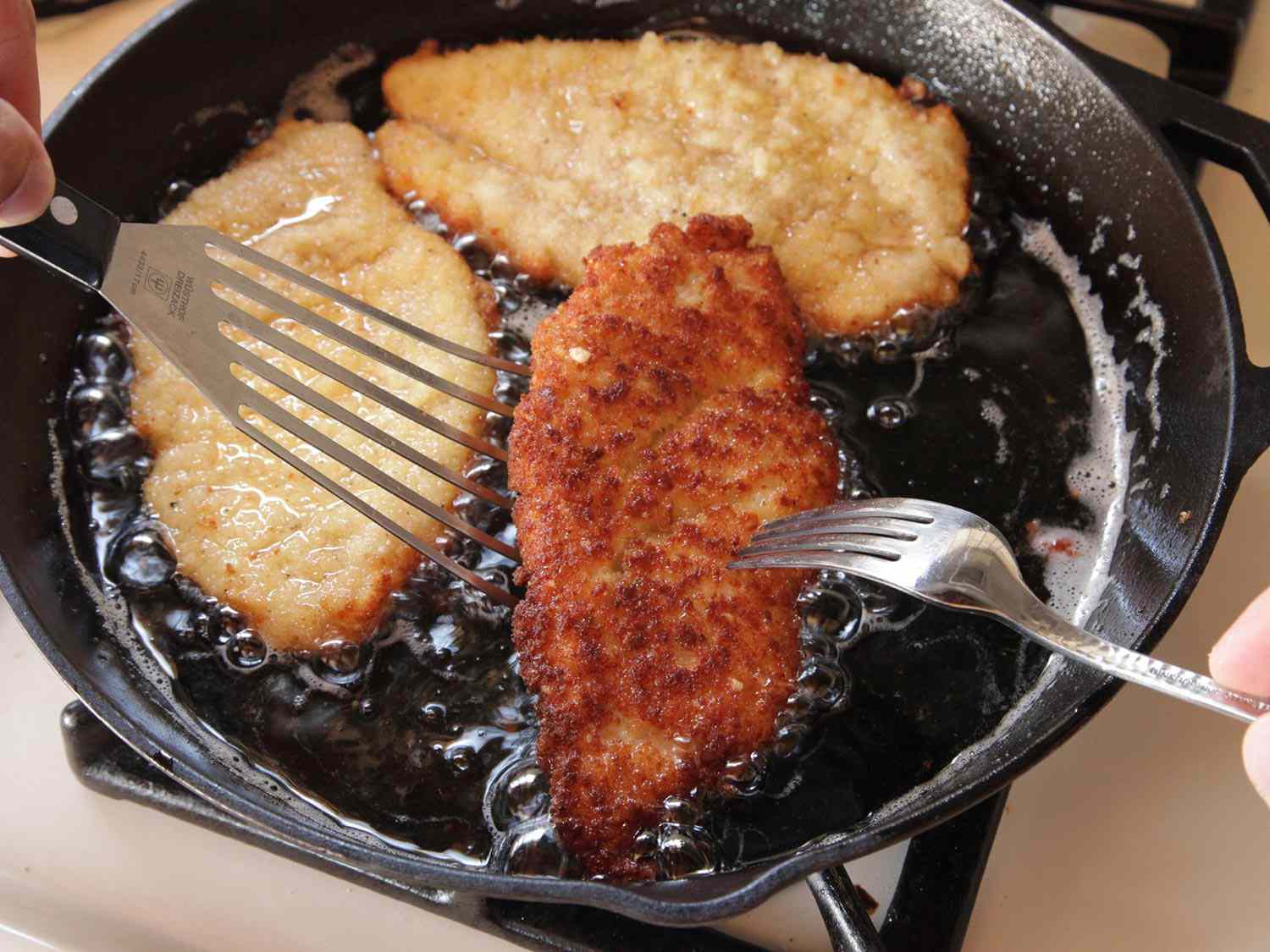 Using spatula and fork to gently flip over golden brown underside fried chicken breast in hot oil