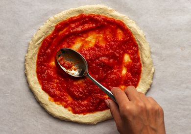 用金属勺将纽约风格的披萨酱抹在生的披萨面团上。