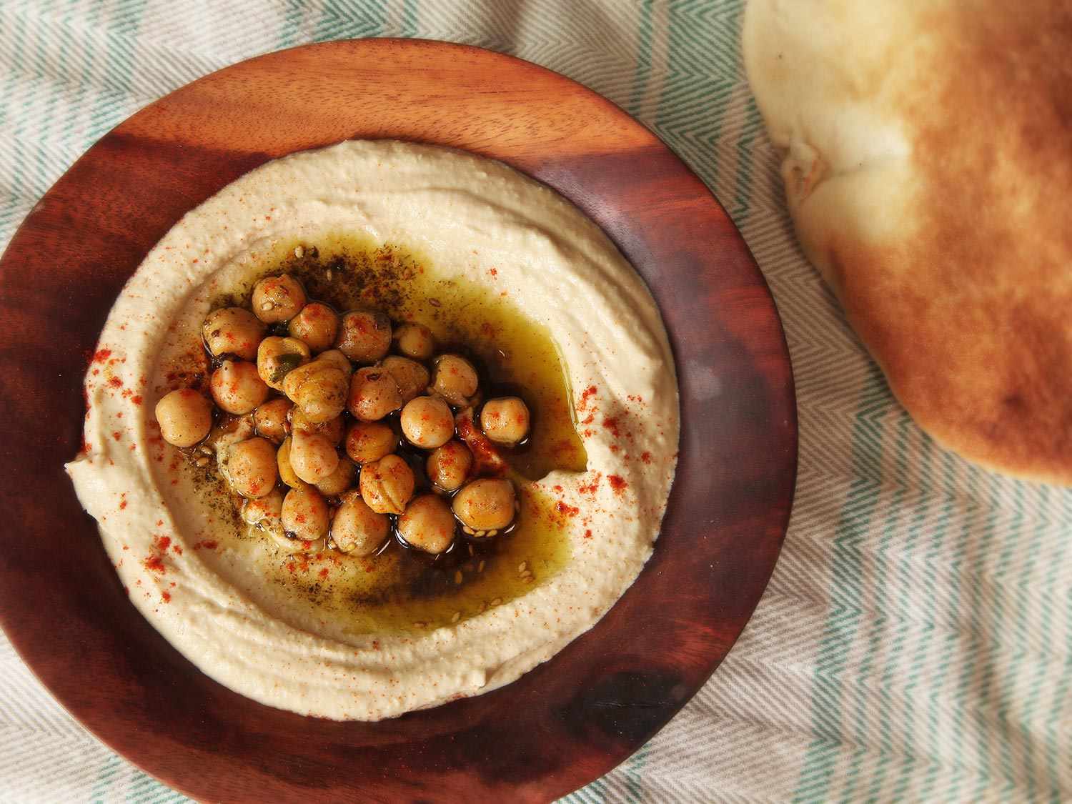 头顶上的一张光滑的以色列式鹰嘴豆泥，上面撒着鹰嘴豆、橄榄油、扎塔尔和辣椒粉，背景是皮塔饼。