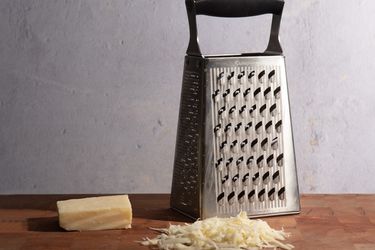 Box grater on a cutting board flanked by a rind of cheese and foregrounded by a pile of shredded cheese
