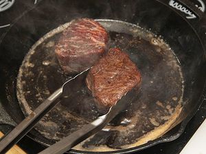 两只小牛排在铸铁煎锅里煮。一把钳子正夹起一块牛排。