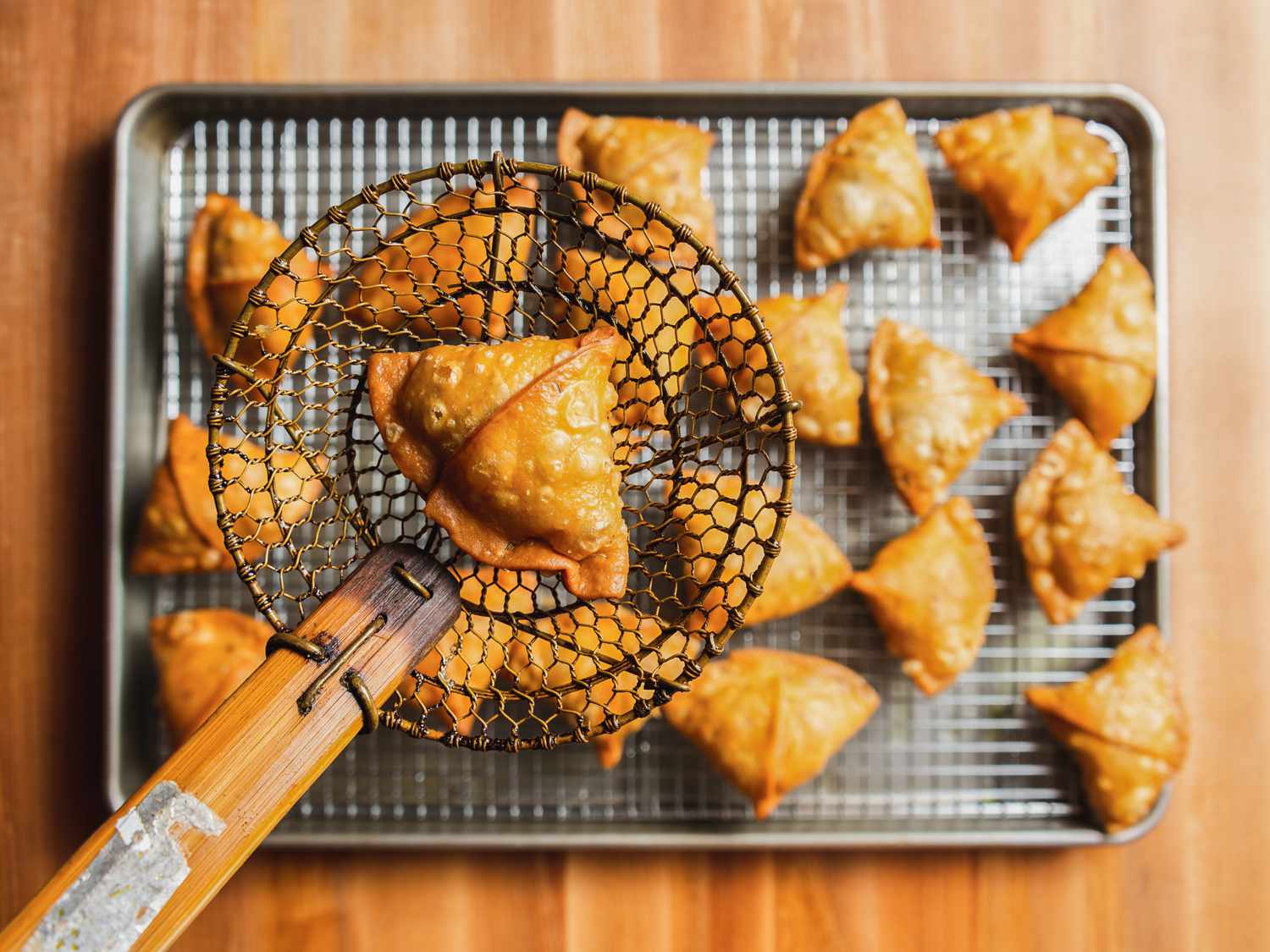 Fried samosa in a spider strainer held aloft a rimmed baking sheet with a wire rack set into it with samosas cooling on top