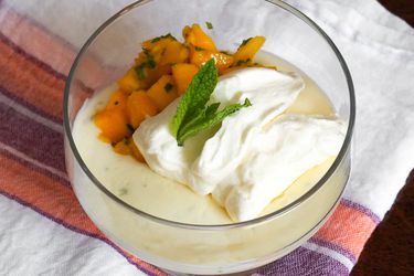 A Lime Posset With Mint and Mango Fruit Salad in a glass dish with mint leaves and whipped cream on top.