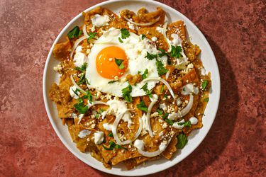 Chilaquiles Verdes topped with a fried egg, on a white plate on a reddish surface.