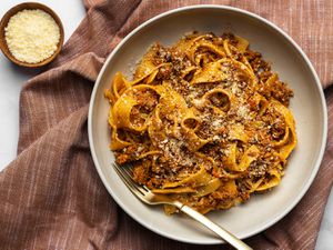 A bowl of pasta with Bolognese sauce on a textured cloth with a small bowl of grated cheese off to the left side.