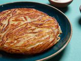 A golden brown potato pancake sits on a plate. It is round, thick, and is made up of clearly defined potato strands.