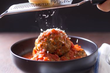 Italian American-style meatballs in a bowl with tomato sauce. Someone is grating parmesan cheese over the meatballs.