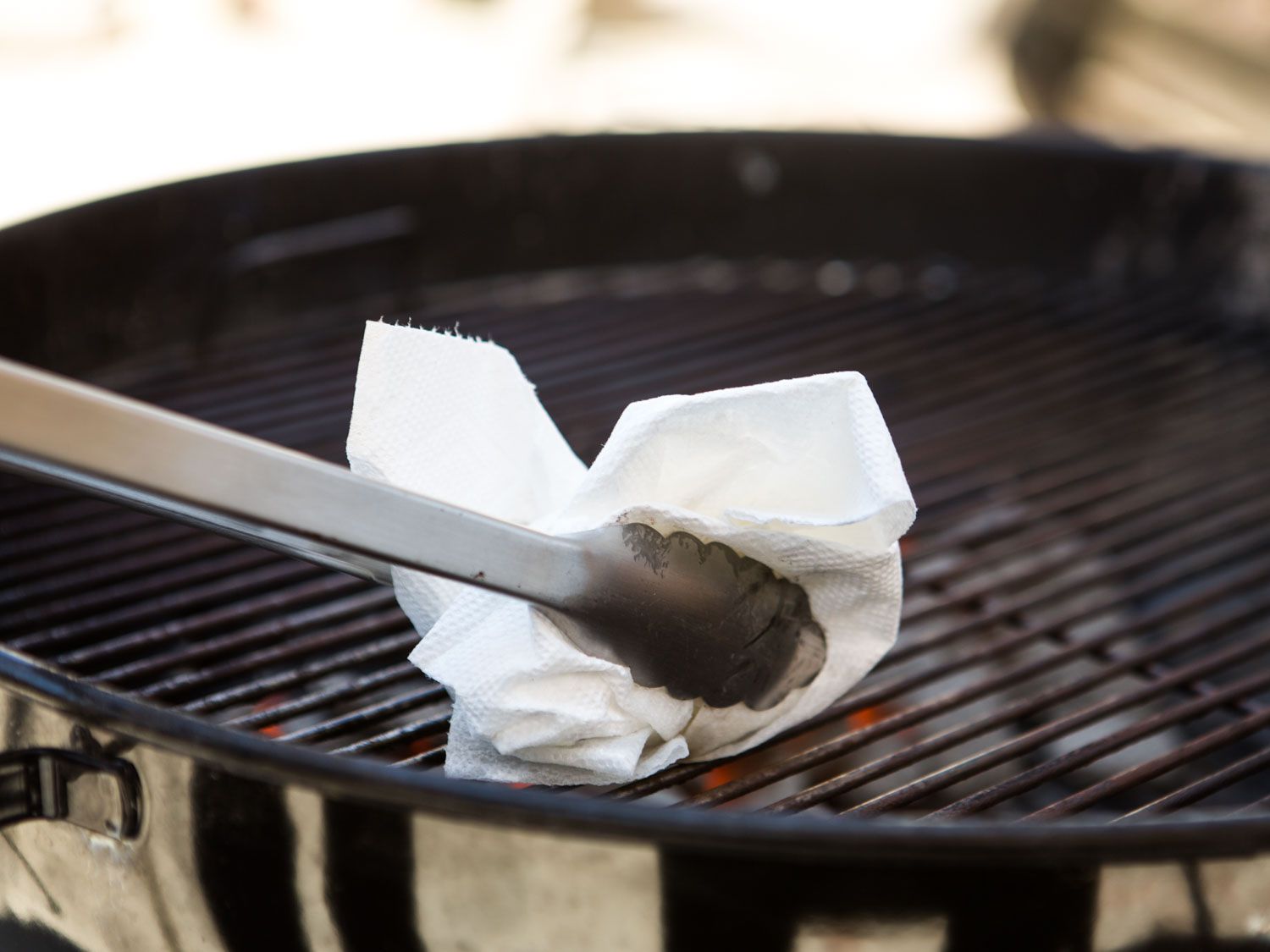 Oiling the grates of a charcoal grill using tongs and a paper towel dipped in neutral oil.