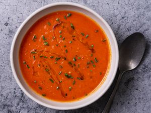 一碗奶油素食番茄汤放在石头背景上，勺子放在碗的右边。汤的表面淋上橄榄油，撒上切碎的香草。