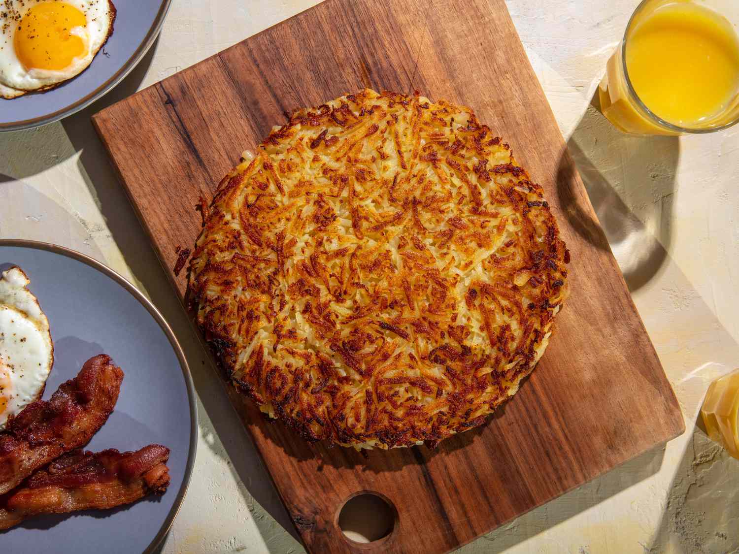 A deeply browned rÃ¶sti sits on a wooden cutting board, with plates of fried eggs and bacon, and glasses of orange juice around it on the table.