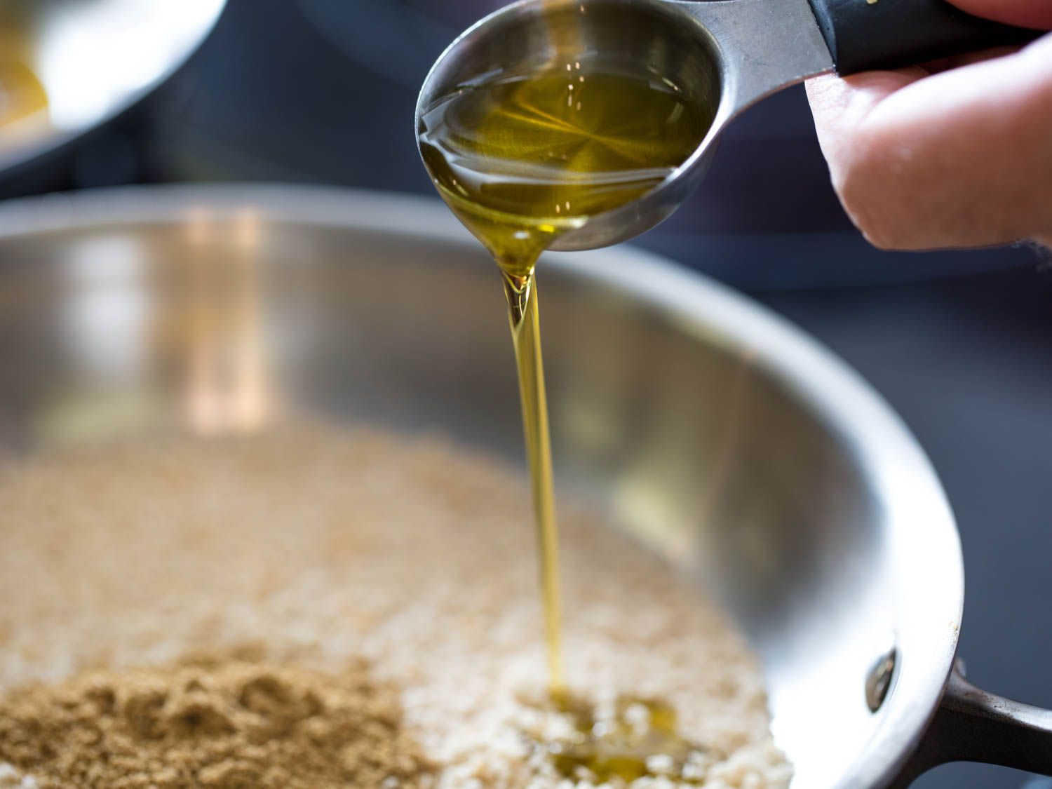 Olive oil being poured into a stainless steel skillet that holds breadcrumbs and ground fennel seeds