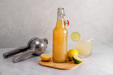 A bottle of sour mix on a wooden cutting board with a lemon, sliced lime, citrus squeezer, and a mixed drink in a glass.