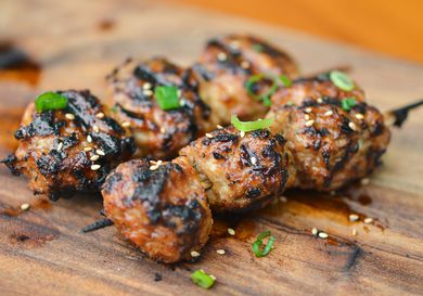 Two skewers of Tsukune, Japanese chicken meatballs, resting on a cutting board and sprinkled with sliced green onions and sesame seeds