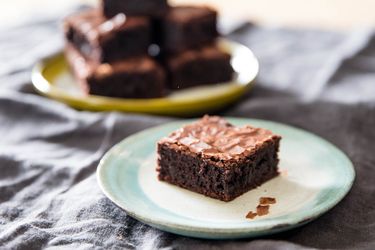 A glossy fudge chocolate brownie on a small plate with stack of brownies in background.