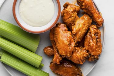 Crispy, sauce-covered fried buffalo wings on a white ceramic plate with sliced celery sticks and a small bowl of bleu cheese dressing, on a white stone background.