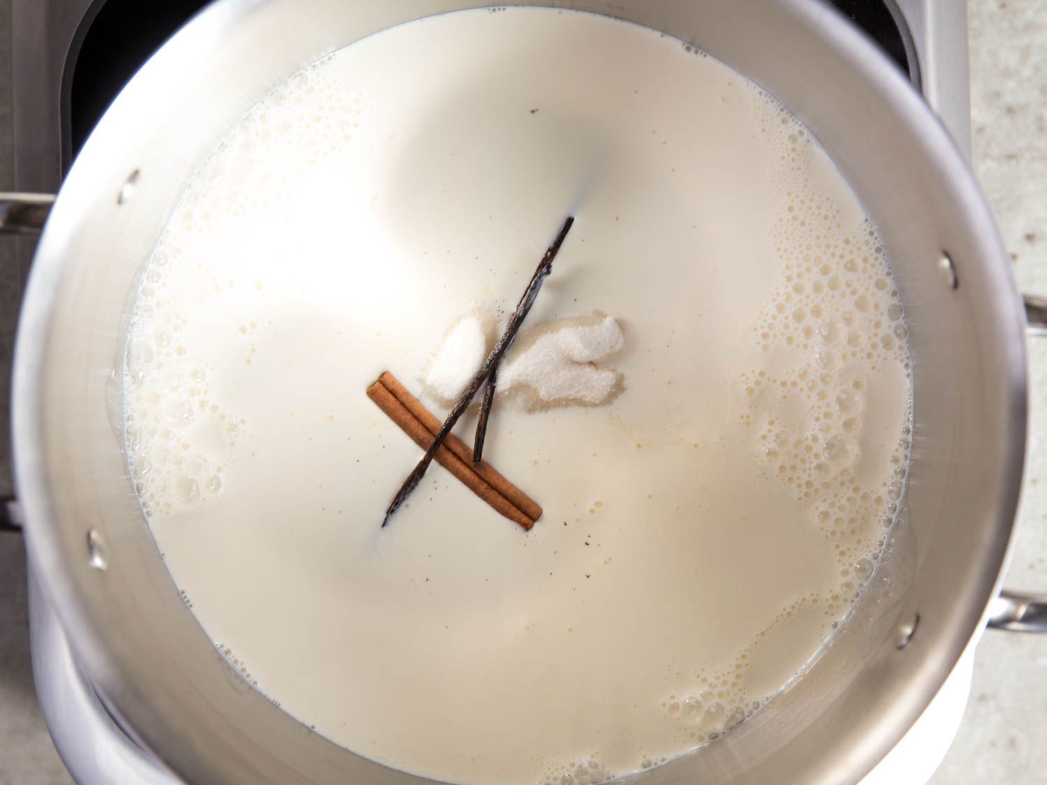 Overhead shot of milk and cream bubbling in a pot, with sugar, a cinnamon stick, and a vanilla bean pod.