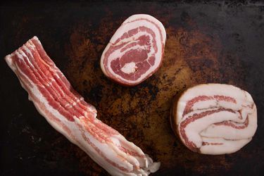 Overhead view of bacon, pancetta, and guanciale arranged on a well-seasoned baking sheet.