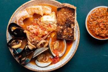A serving of cioppino in a bowl, overflowing with plump shrimp, mussels, clams, calamari, fish, and more. There's a piece of deeply toasted sourdough on the side, and a small bowl holding a roasted red pepper condiment.