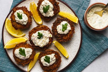 Six crispy herbed crab cakes, each topped with tartar sauce and a single leave of flat parsley, interspersed with slices of lemon. The plate is on a blue textured cloth, and there's a small bowl of tartar sauce and a spoon in the top right corner of the image.