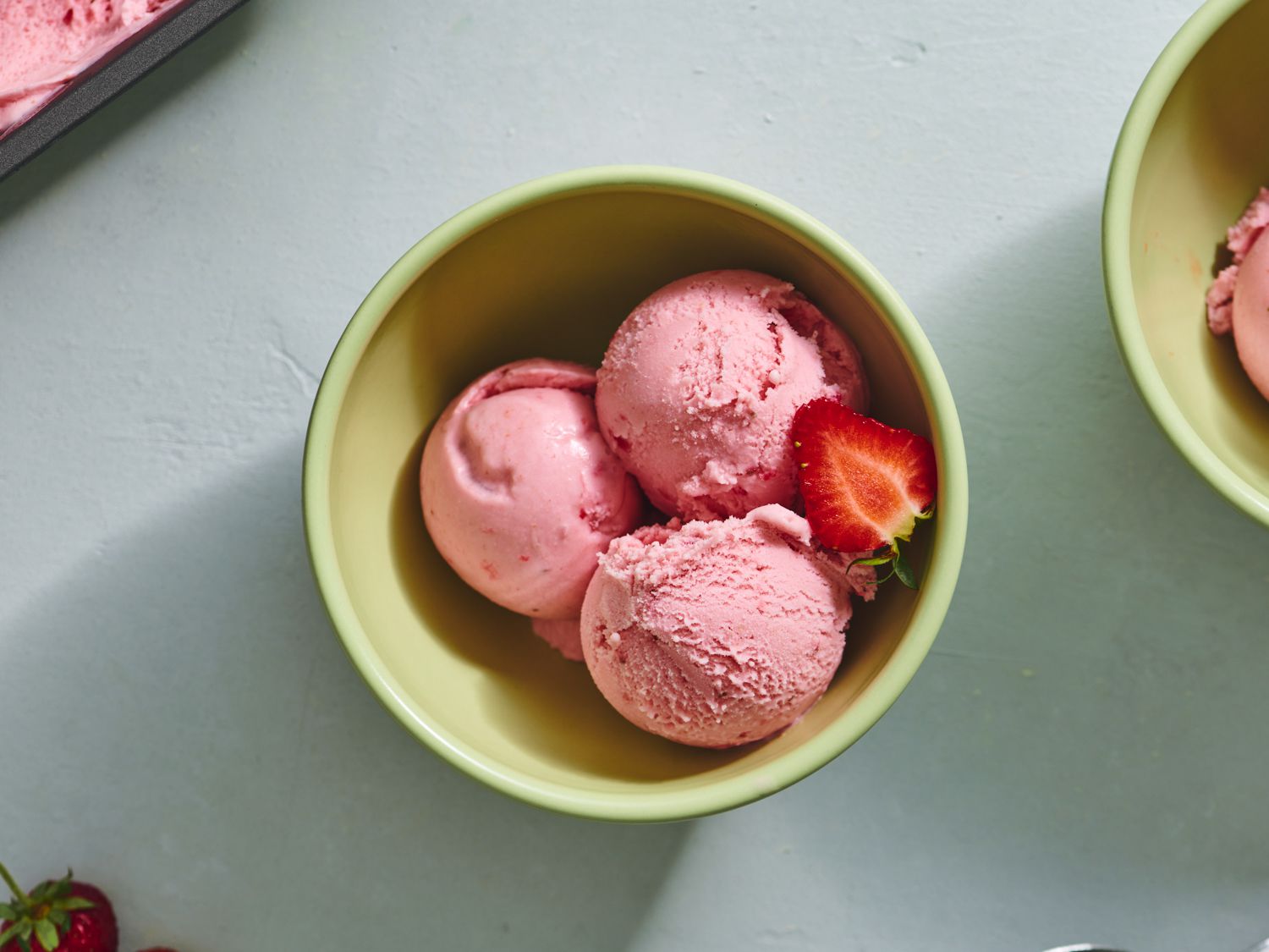Three scoops of strawberry ice cream in a ceramic bowl, with half of a fresh strawberry.