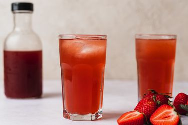 两杯浆果灌木。在图片的右侧是一些草莓，有完整的草莓，也有切成两半的草莓，而在图片左侧的背景中，是一个小玻璃瓶，里面装着更多的灌木。