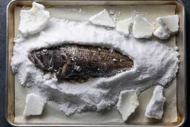 A whole roasted black bass that was buried in a mound of salt on a baking sheet is exposed after the salt crust was cracked and removed.