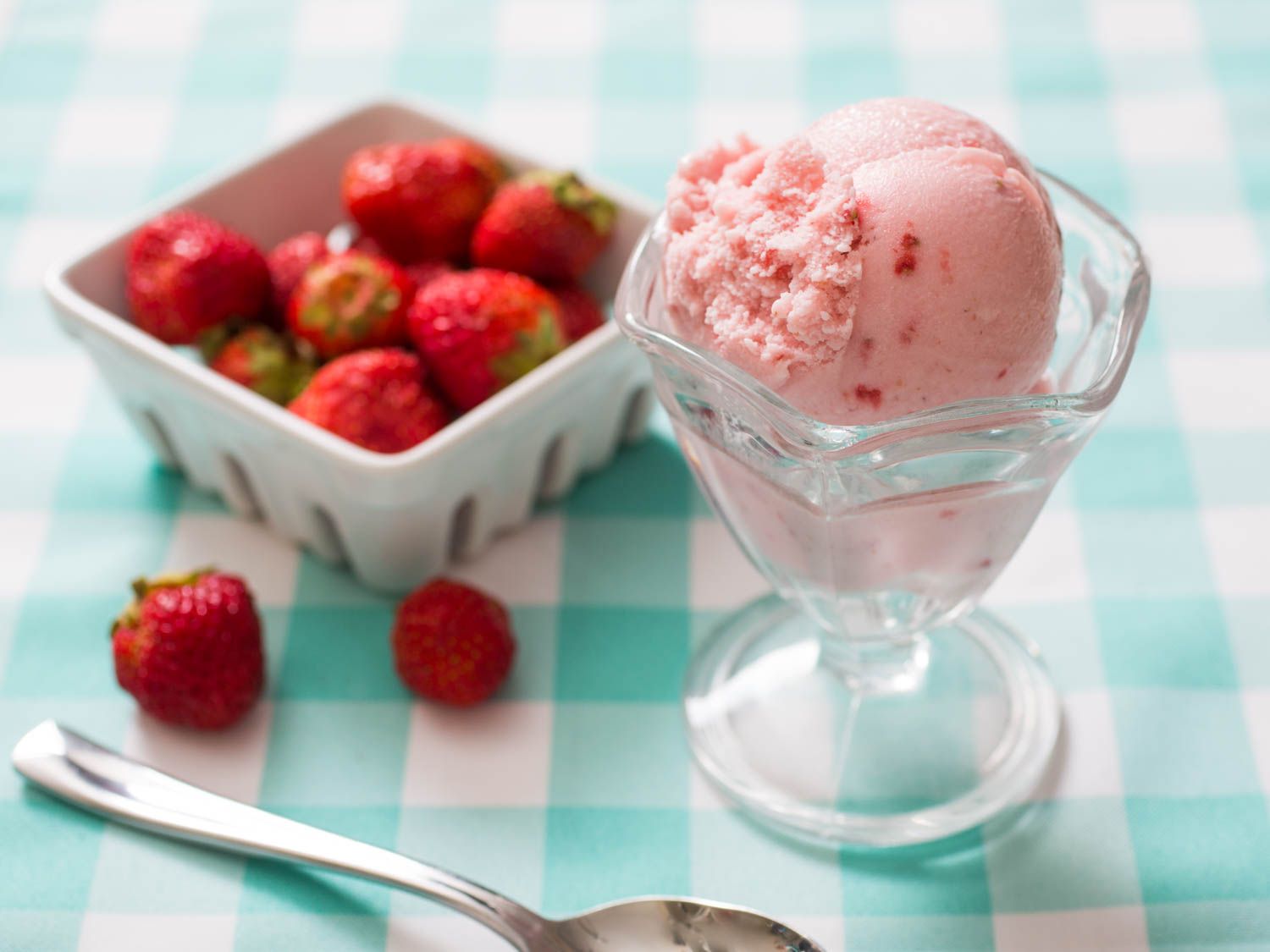 A scoop of strawberry ice cream in a footed glass dish next to a container of strawberries.