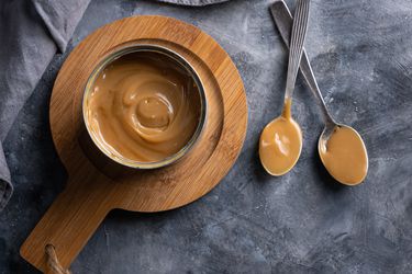 A can of dulce de leche on a wooden trivet with two metal spoons with dulce de leche on them, on a stone background