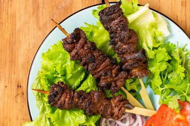 A plate with three Nigerian beef suya (spiced grilled skewers) on a bed of lettuce with sliced red onions, tomato sliced, and cilantro.