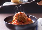 Grating Parmesan cheese on top of Italian-American meatballs in red sauce in a dark bowl.