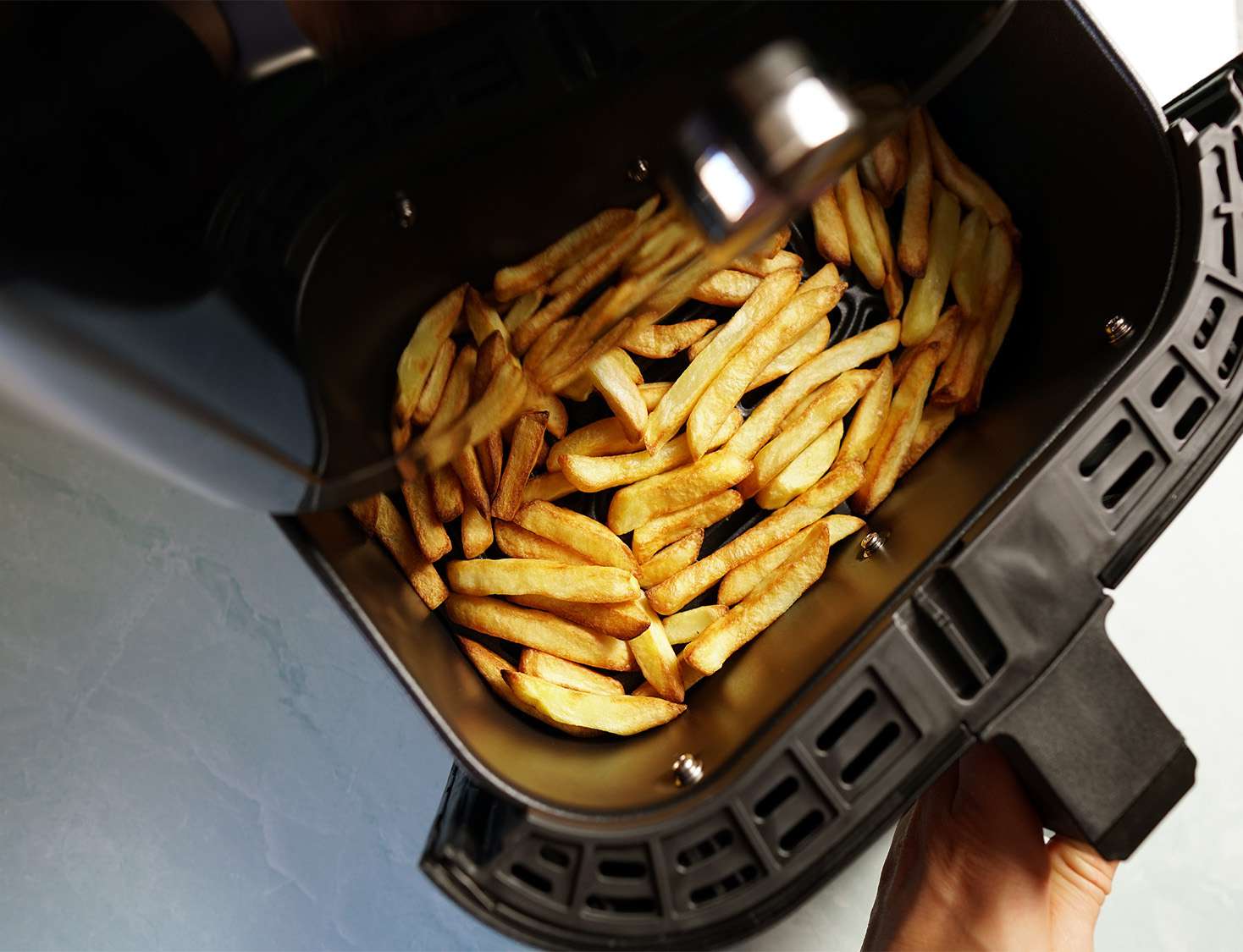 cooked fries in an air fryer basket with a hand pulling the basket out midway