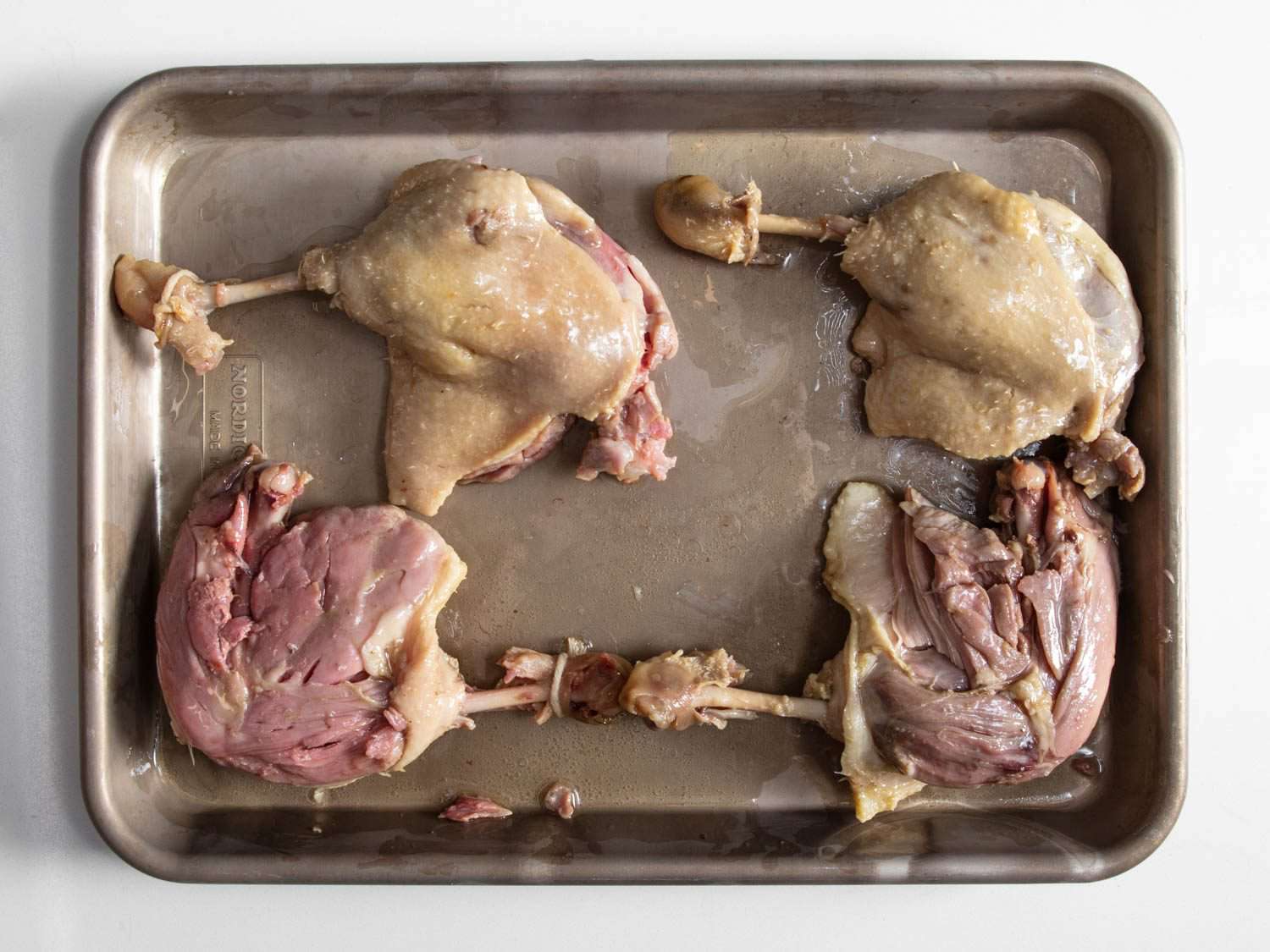 Overhead comparing duck confit made with legs cured with pink salt and kosher salt, and legs cured with just kosher salt. The meat on legs cured with pink salt is rosy, while salt-cured legs are brown.