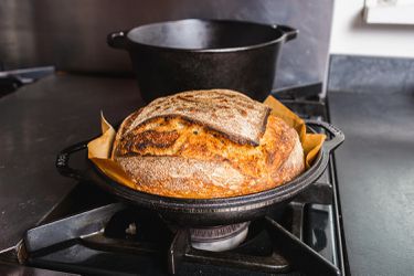 A round bread loaf in a parchment paper sling sitting in the shallow base of a cast iron combo cooker
