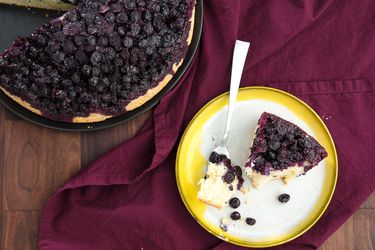 A plated wedge of upside down blueberry muffin cut from a whole cake, with a fork pierced into a piece,