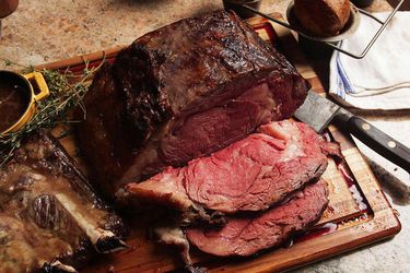 A whole cooked prime rib roast with two slices on a wooden cutting board with a knife, herbs, and sauce.