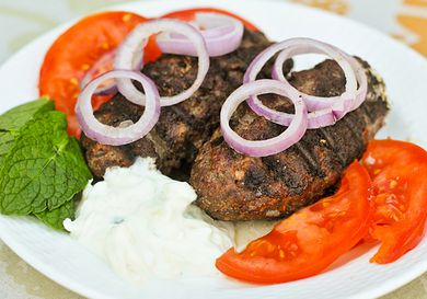 Greek bifteki on a plate with tomato slices, red onion slices, mint leaves, and tzatziki.