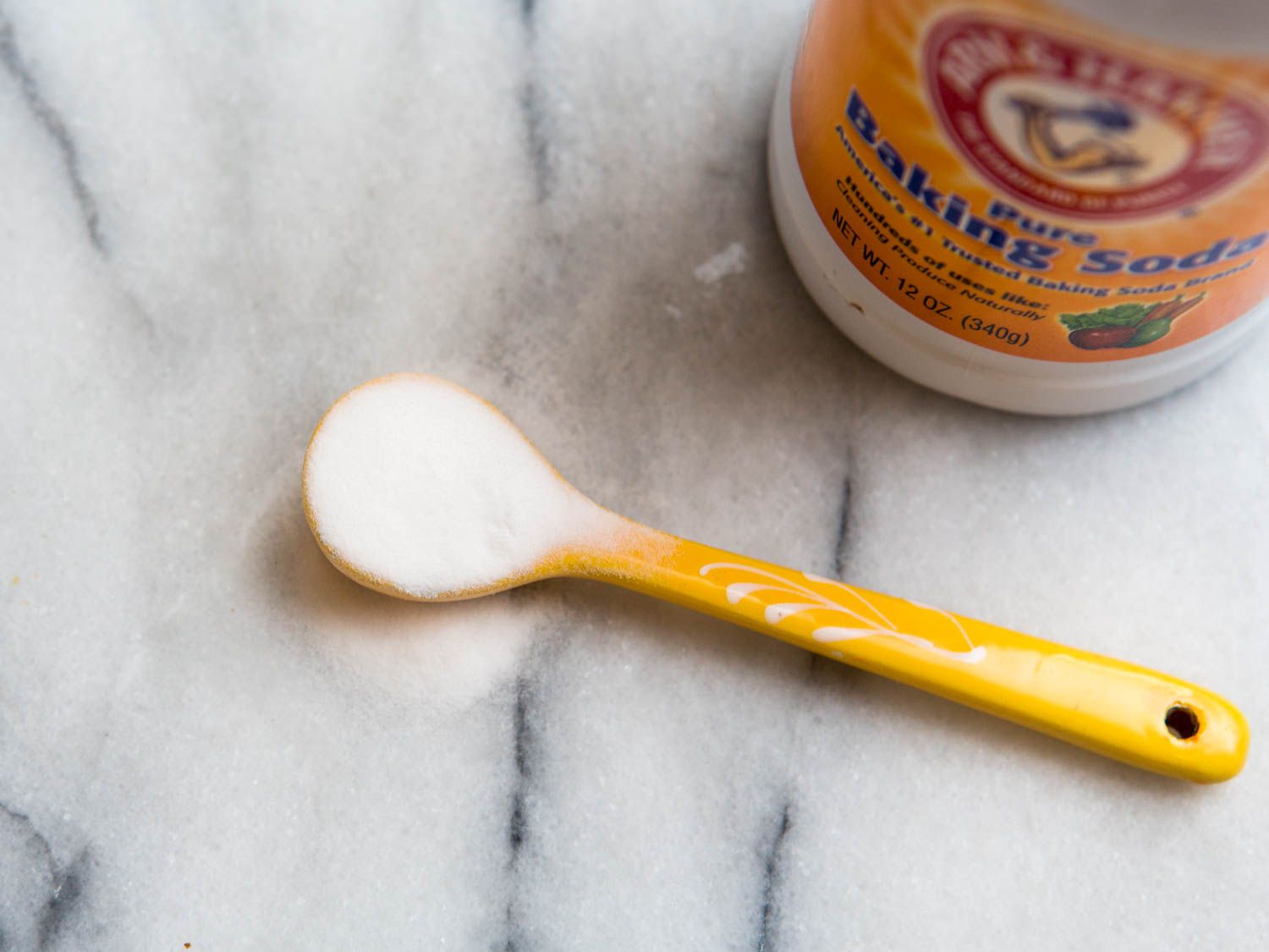 A yellow spoon holding baking soda with a container of baking soda next to it.